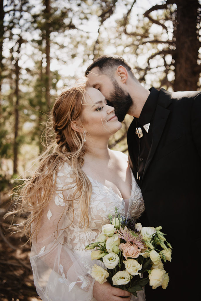 groom leaning down to kiss his bride on the cheek as she looks up to him and smiles in the woods captured by jackson hole photographers 