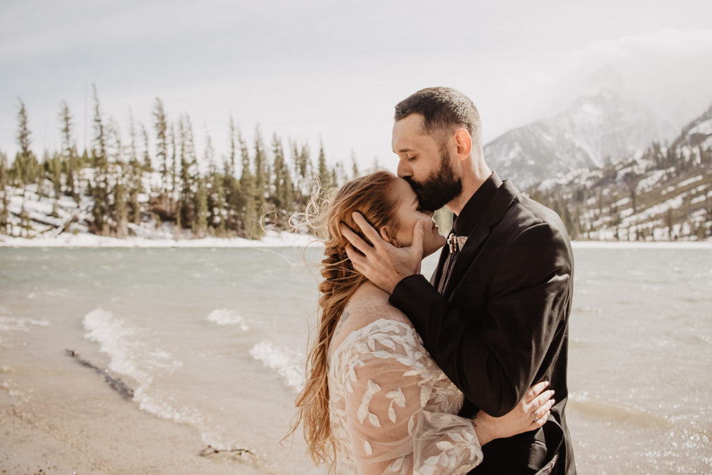 jackson hole elopement with bride adn groom standing next to a lake in the grand tetons with groom holding brides head and kissing her forehead as she smiles with her eyes closed with the water in the distance and beyond that the forest
