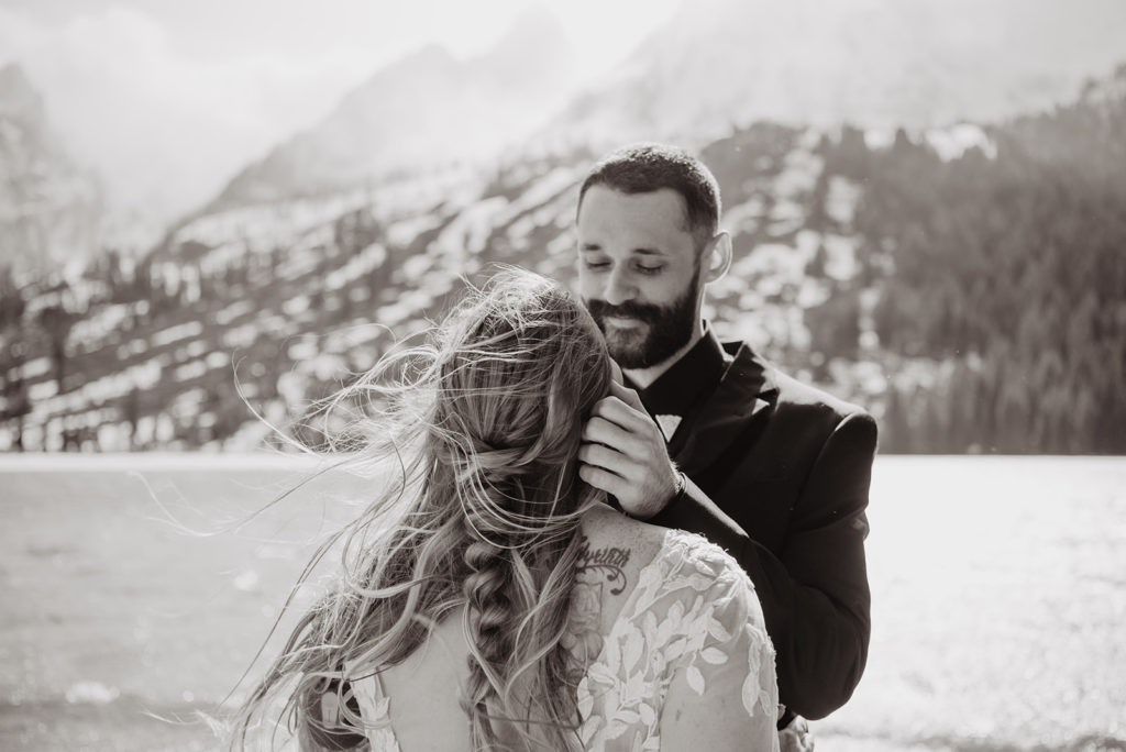 groom smiling at his bride as he caresses her cheeks with the Tetons behind them and a lake
