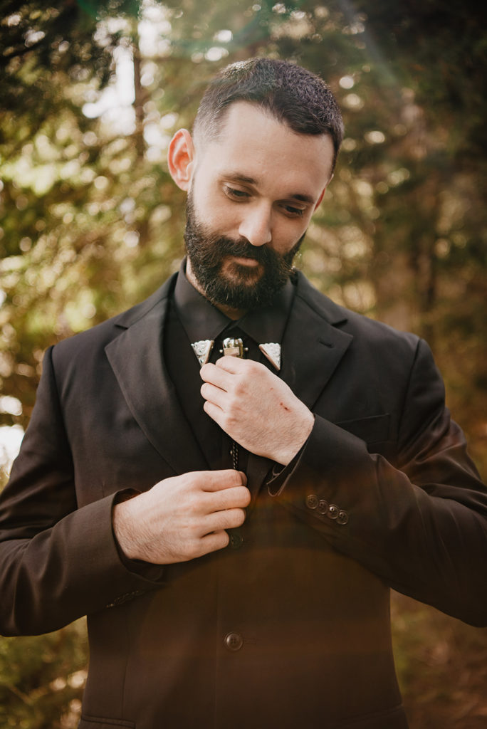 groom fixing his bolo tie and looking downwards in the woods 