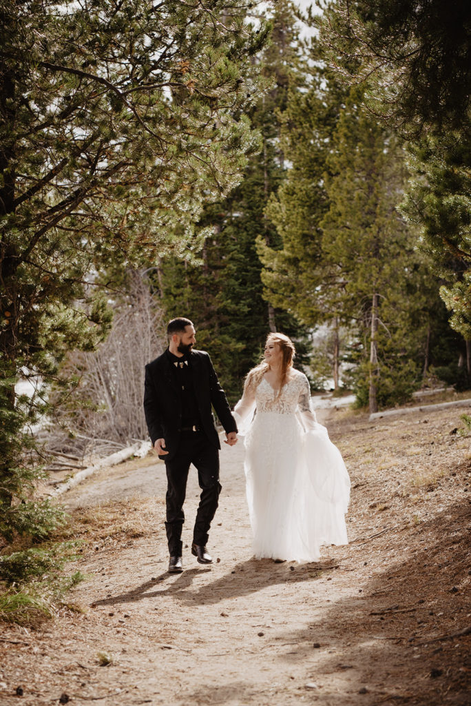 bride and groom holding hands and walking through the woods in the Tetons while the sun shines through the trees onto them captured by Jackson hole photographers 
