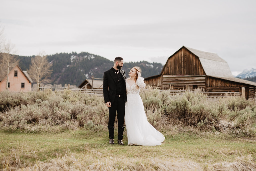 Jackson Hole photographers capture bride and groom standing outside of an old barn at Mormon Row on a cloudy and romantic wedding day 