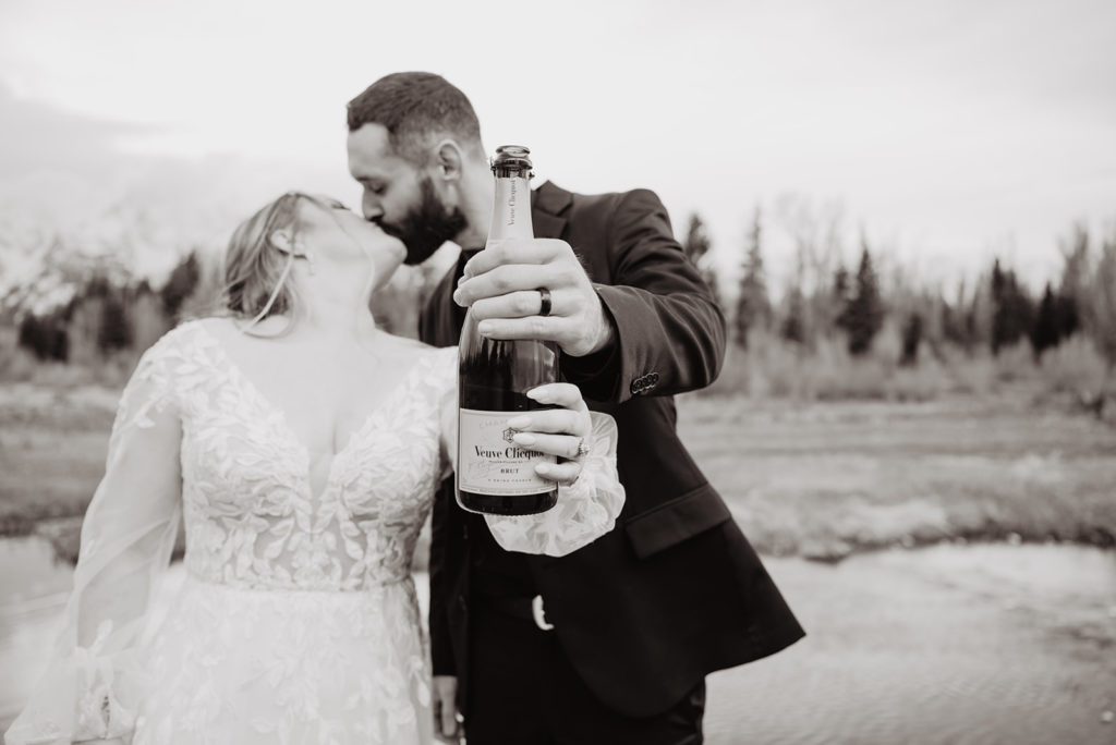 bride and groom holding a bottle of champagne together while kissing in celebration