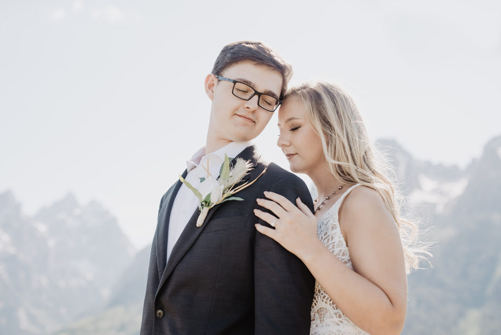 bride standing behind her groom as she holds his shoulder and leans into his while the groom looks back at her with the mountains of the Grand Tetons behind them 