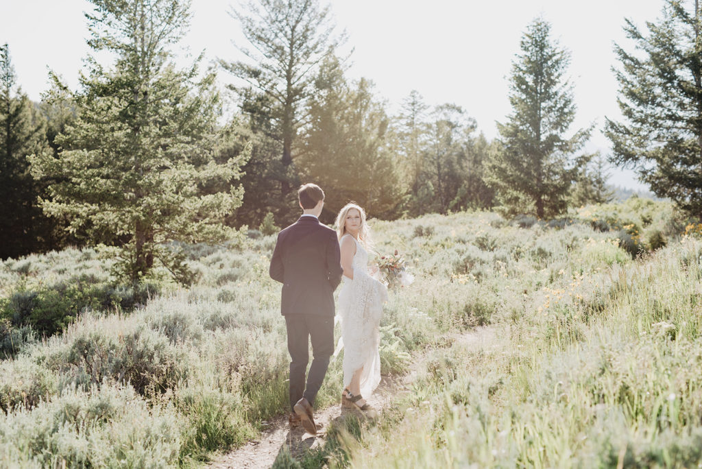 Jackson Hole wedding photographer photographs bride adn groom walking together through the woods of the Tetons with the bride ahead of the groom and looking back to him