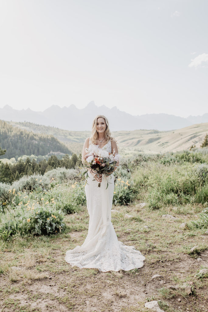 bride standing in a field in Jackson Hole with the mountains in the distance as she holds her wedding florals and smiles captured by Jackson Hole wedding photographer