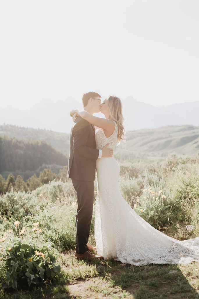 bride holding her arms around the groom's shoulders as the groom holds her waist and they kiss while the sun shines over them in Jackson Hole