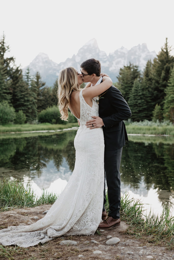 bride adn groom embracing and kissing on a lake in the Grand Tetons with the forest behind the lake and the mountains further in the distance captured by Jackson Hole wedding photographer