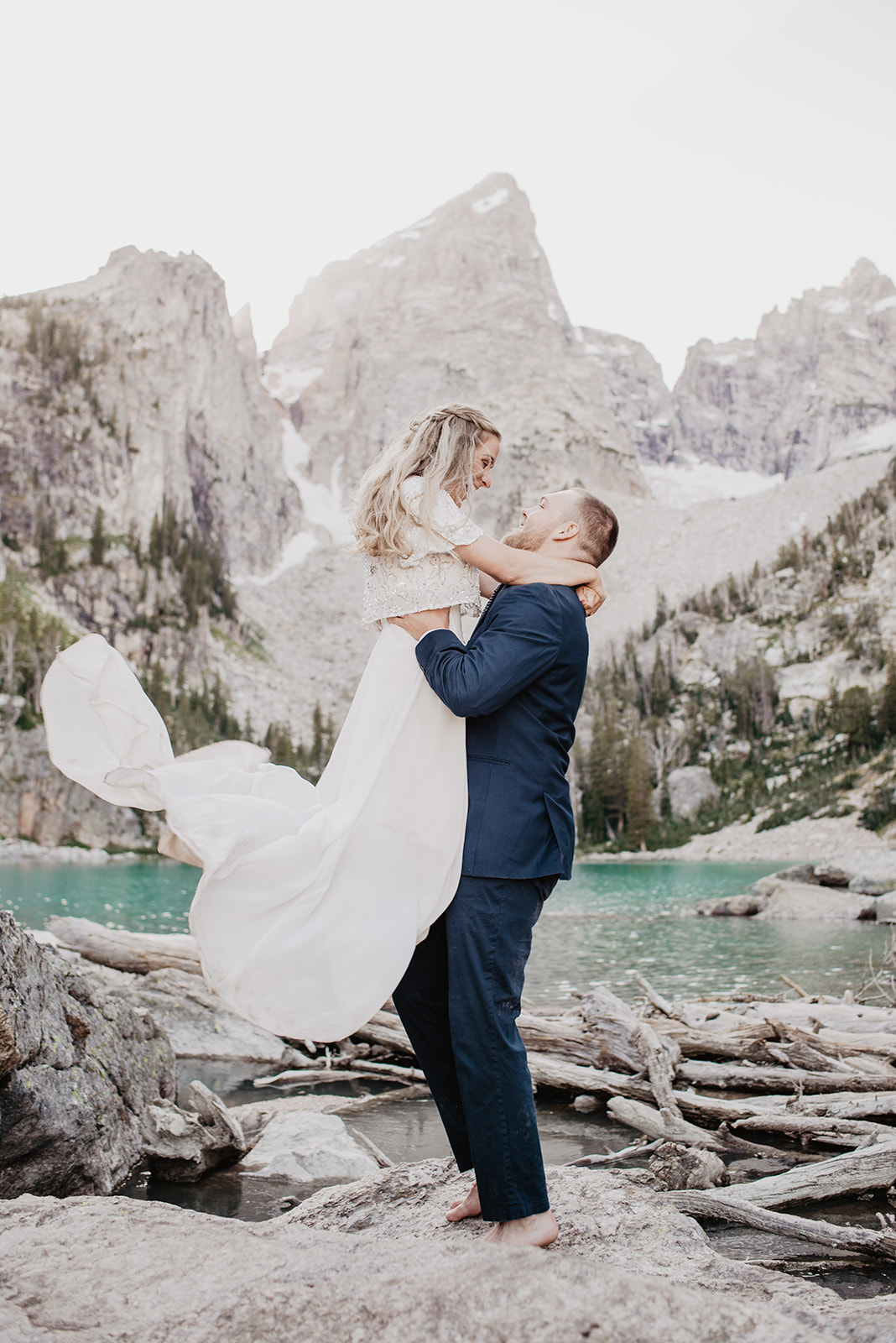groom lifting his bride up in the air as her wedding dress flows behind her with the Tetons and a lake being the couples photographed by Jackson Hole wedding photographers