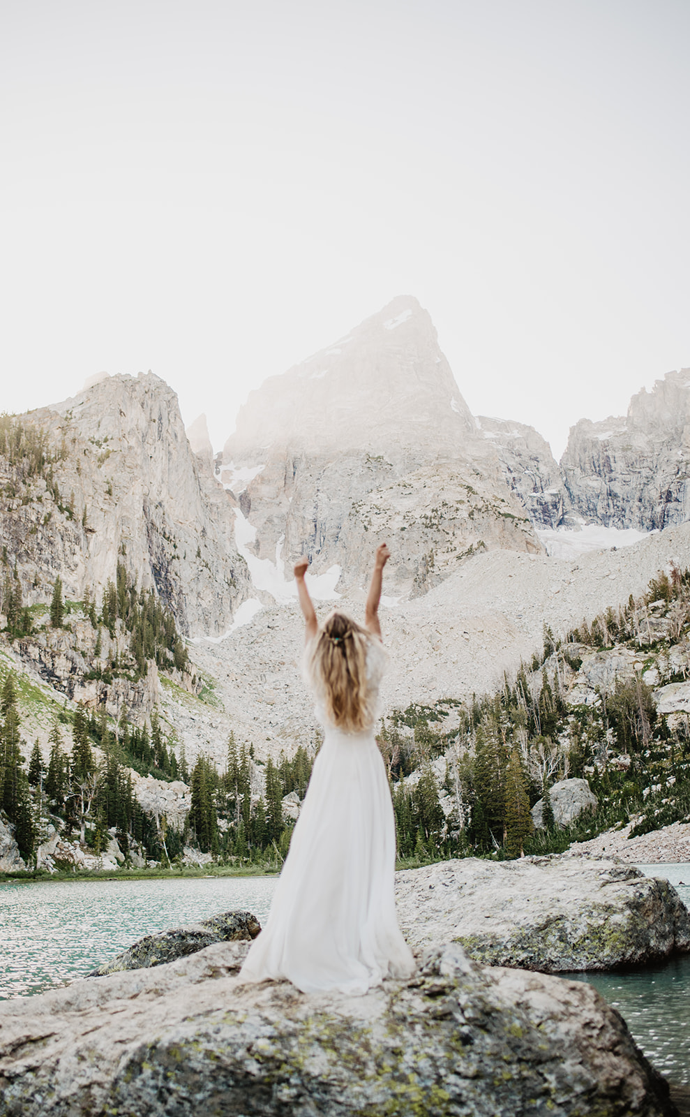 bride in the Tetons for her Jackson Hole wedding with her hands raised in the air in celebration with a blue lake in front of her captured by Jackson Hole wedding photographer