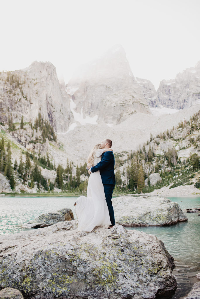 bride hugging her groom as he picks her up on a rock in the tetons with a bright blue lae behind them for their wyoming elopement pictures with jackson hole photographers 