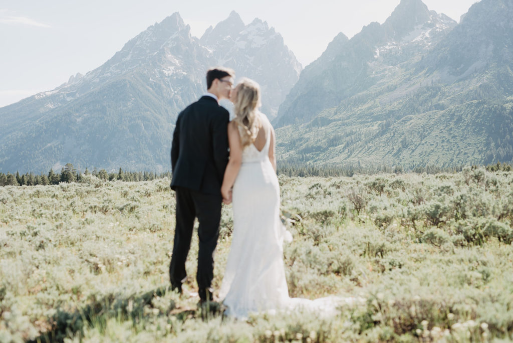 bride and groom kissing in a meadow in the Tetons with the sun shining over the mountains before it sets behind them for wyoming wedding 