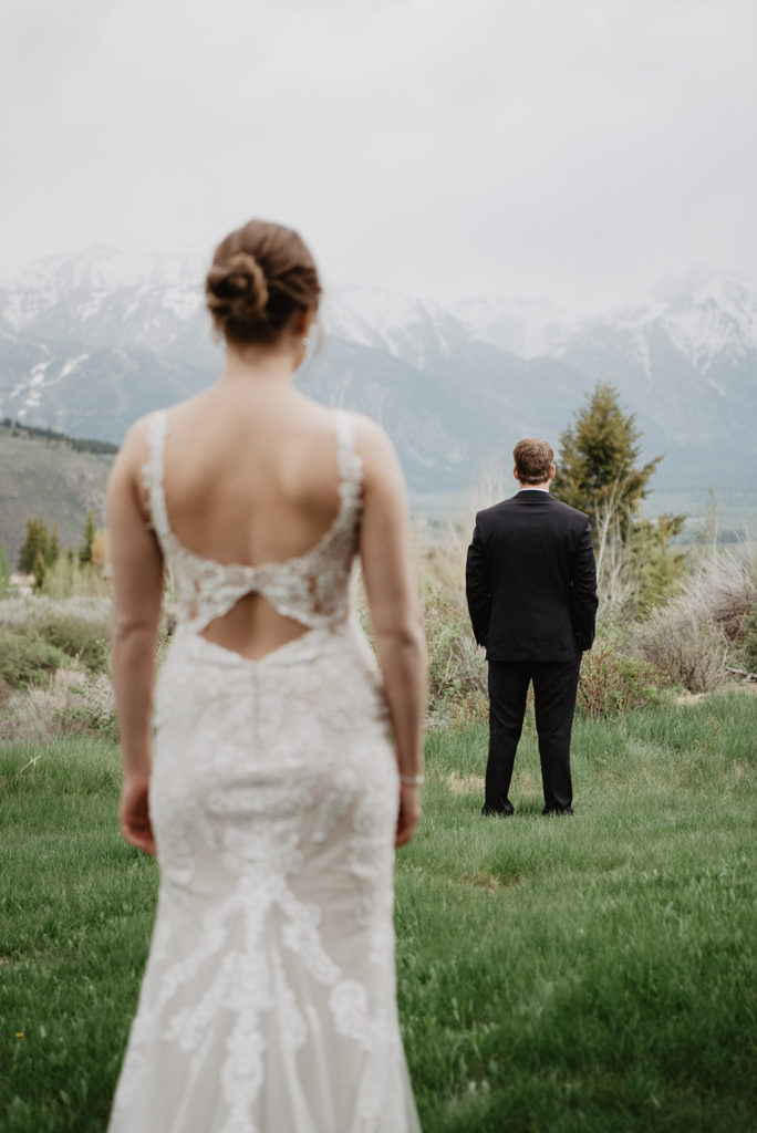 bride in a lace wedding dress walking towards her groom as he faces the other way for their wedding first look at their Jackson Hole wedding int he mountains with the Tetons in the distance 