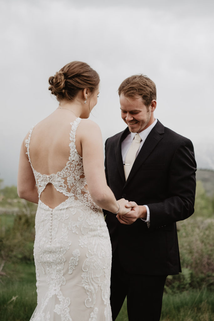 groom holding his brides hands while looking at her dress for their first look together documented by Jackson Hole wedding photographers 