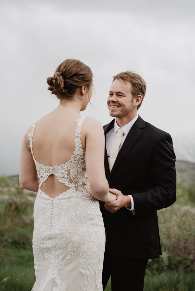 groom holding his brides hands during their first look for their Jackson Hole wedding in the Tetons with the groom smiling at his bride as she wears a lace wedding dress
