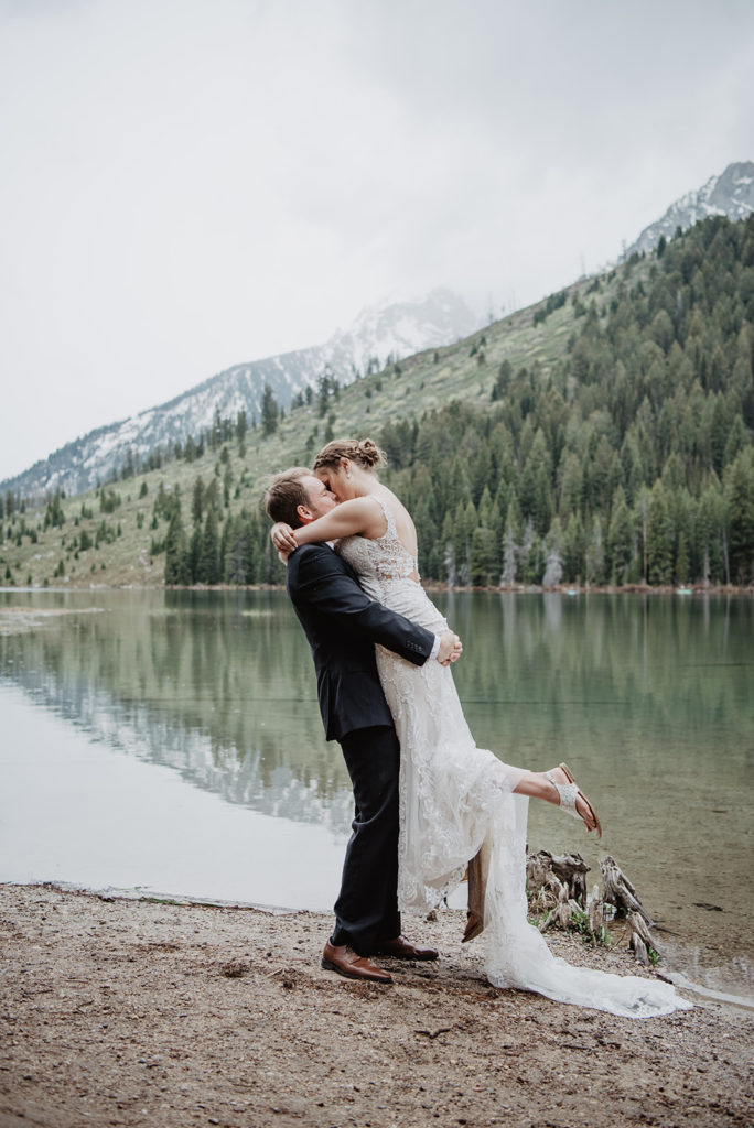 groom picking up his bride on the beach of a lake in the Tetons as her lace elopement dress trails behind her captured by Jackson Hole photographer