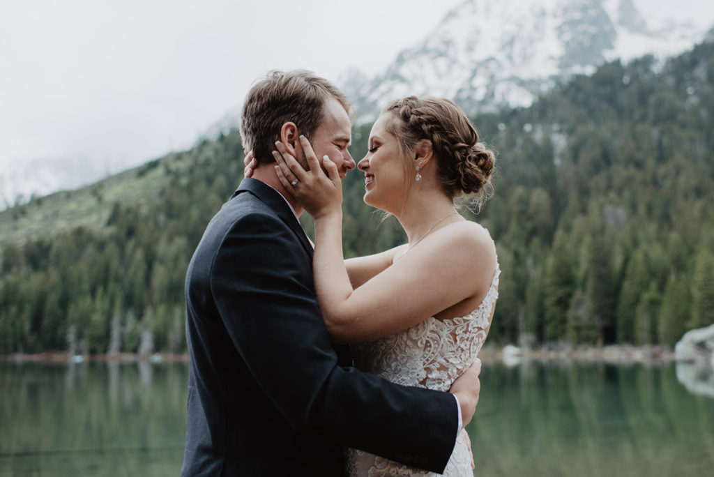 bride holding her grooms face and smiling as he holds her around the waist with a lake, treeline and mountains in the distance 