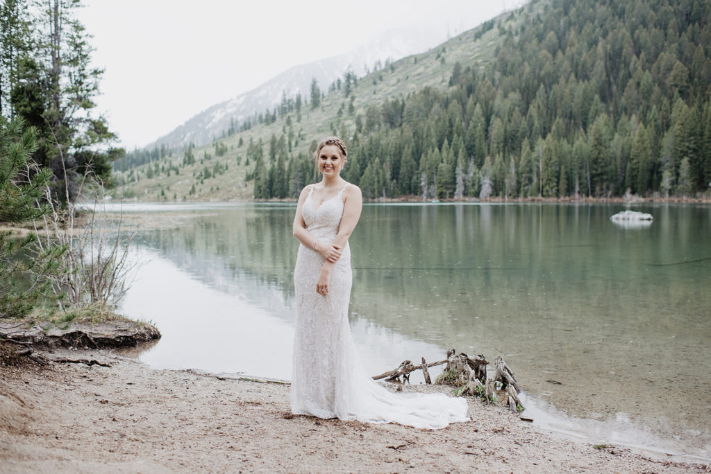 jackson hole photographers captures bride standing next to string lake in her bridal gown with her lace train spread on the sand for her grand teton wedding 