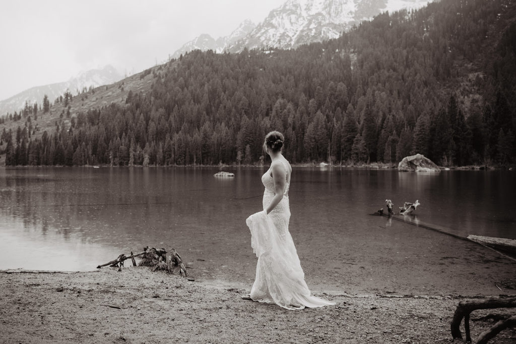 Jackson Hole wedding photographer captures bride in her lace wedding dress walking along a lake in the Tetons with the mountains in the distance for Jackson Hole wedding 