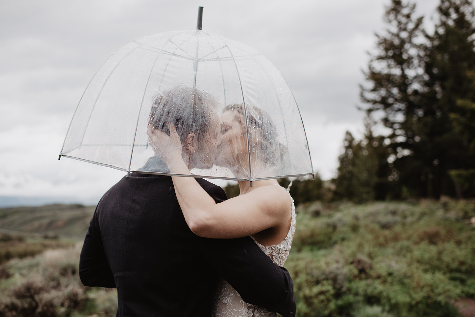 rainy wedding day with bride adn groom under a clear umbrella kissing as the bride embraces her groom captured by Jackson Hole wedding photographers