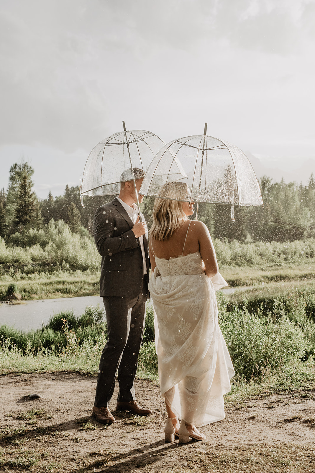 Jackson Hole photographer with bride and groom under a clear umbrella while it rains around them and they look into the sunset together for their Grand Teton wedding