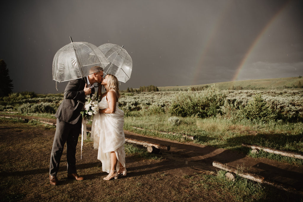 dark stormy sky with two rainbows in the distance with bride and groom kissing under clear umbrellas overhead for rainy day wedding