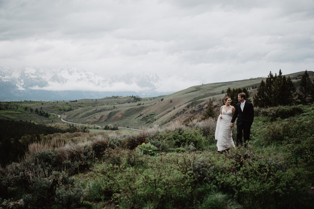 bride and groom holding hands and walking through a field in the Tetons that looks over a valley with Jackson Hole wedding photographer 