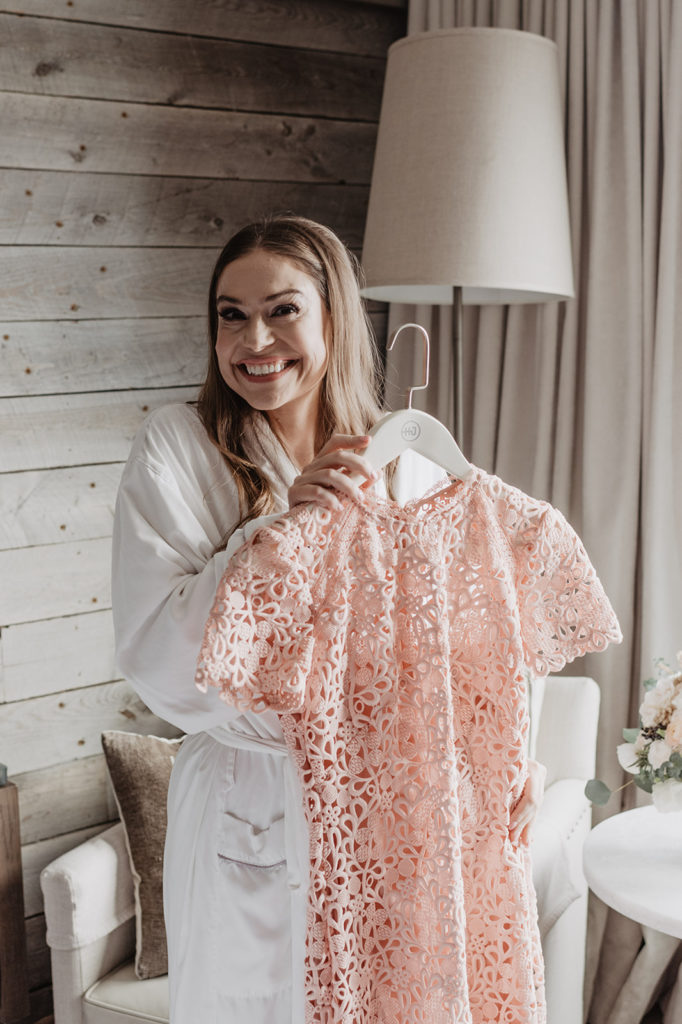 bride getting ready in a white robe as she holds up her pink wedding dress and smiles photographed by Jackson Hole wedding photographer