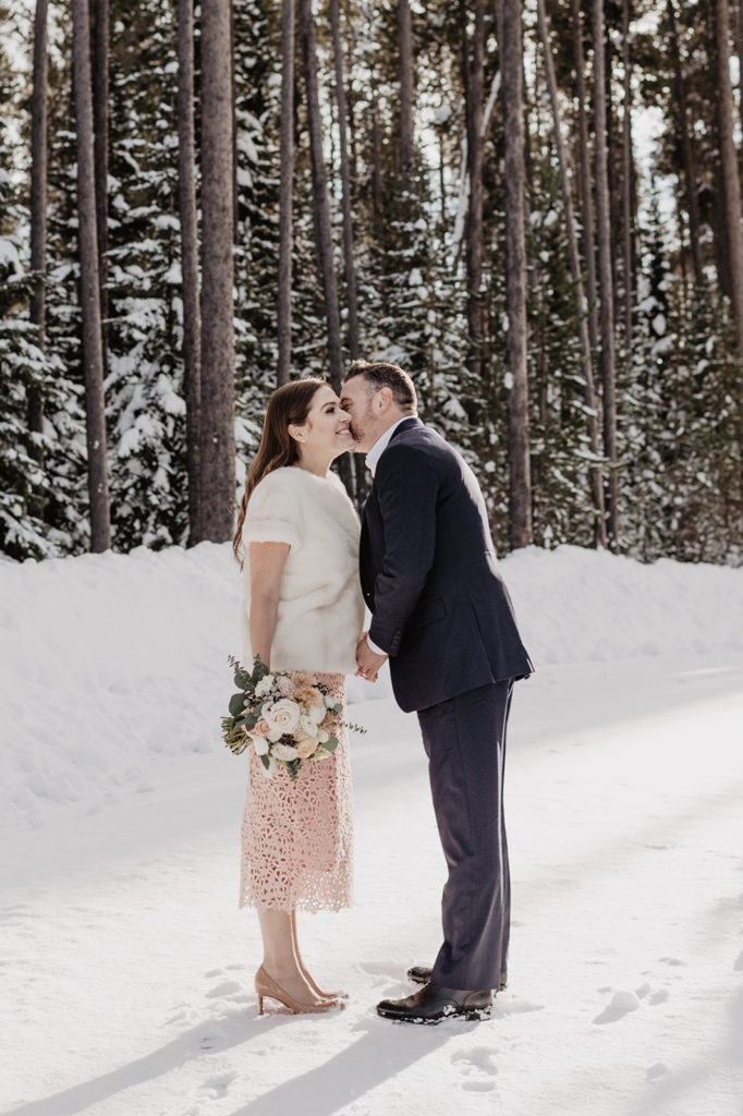 Jackson Hole wedding photographer captures bride and groom in the woods together with groom kissing his bride on the cheek as she smiles 