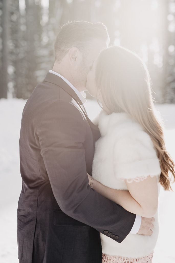 sun shining through pine trees as the bride and groom kiss each other romantically for their snow winter wedding with Jackson Hole wedding photographer 