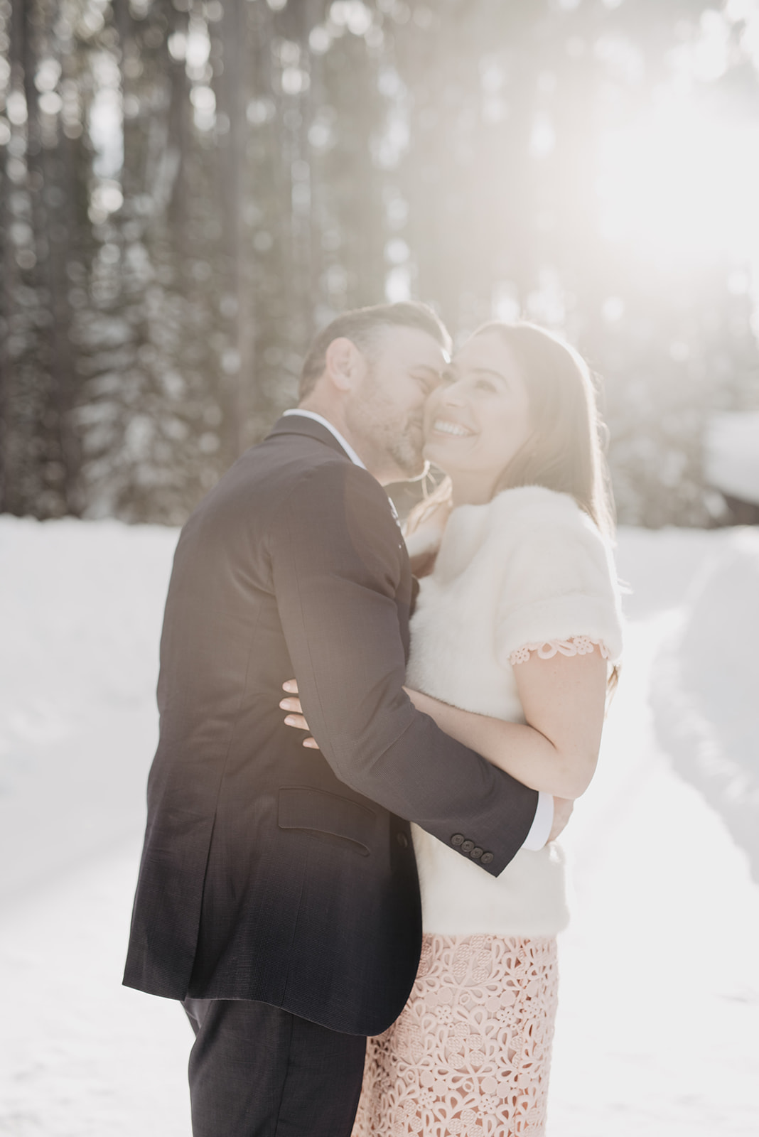 groom kissing brides cheek as she holds onto him and smiles with the sun shining behind them through the pine trees int he Tetons by Jackson Hole wedding photographer