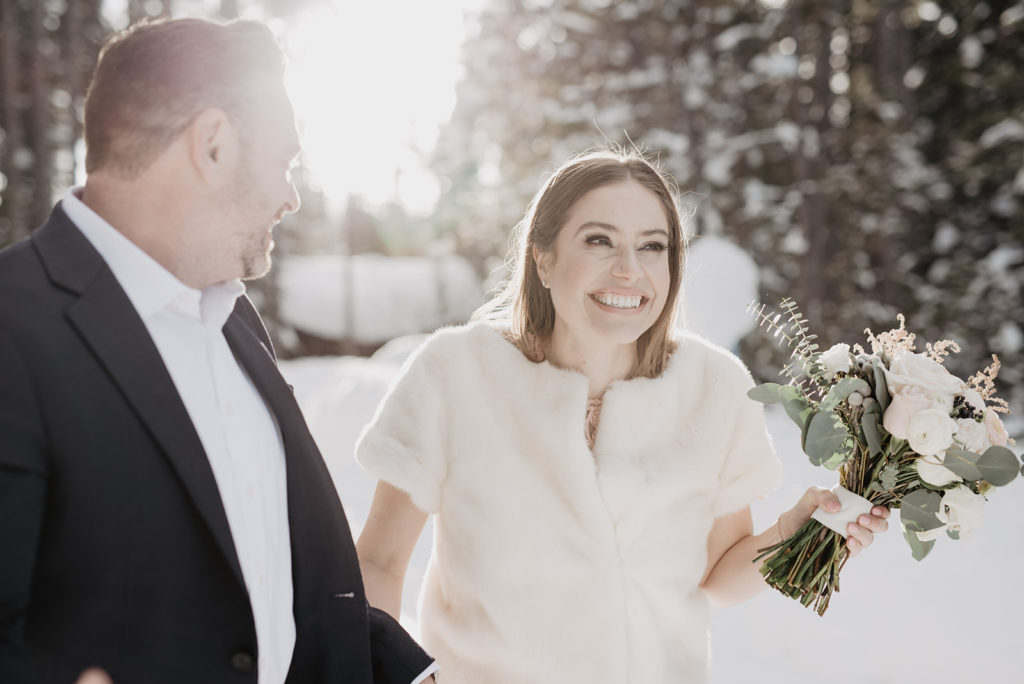 candid wedding photo of bride and groom holding hands and smiling as they walk through the woods together while the sun shines behind them on their winter wedding captured by Jackson Hole wedding photographer