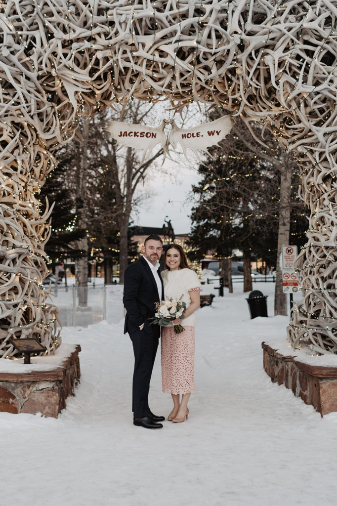 bride and groom standing under the iconic antler arch in Jackson Hole and smiling with snow surrounding them photographed b yJackson Hole wedding photographer