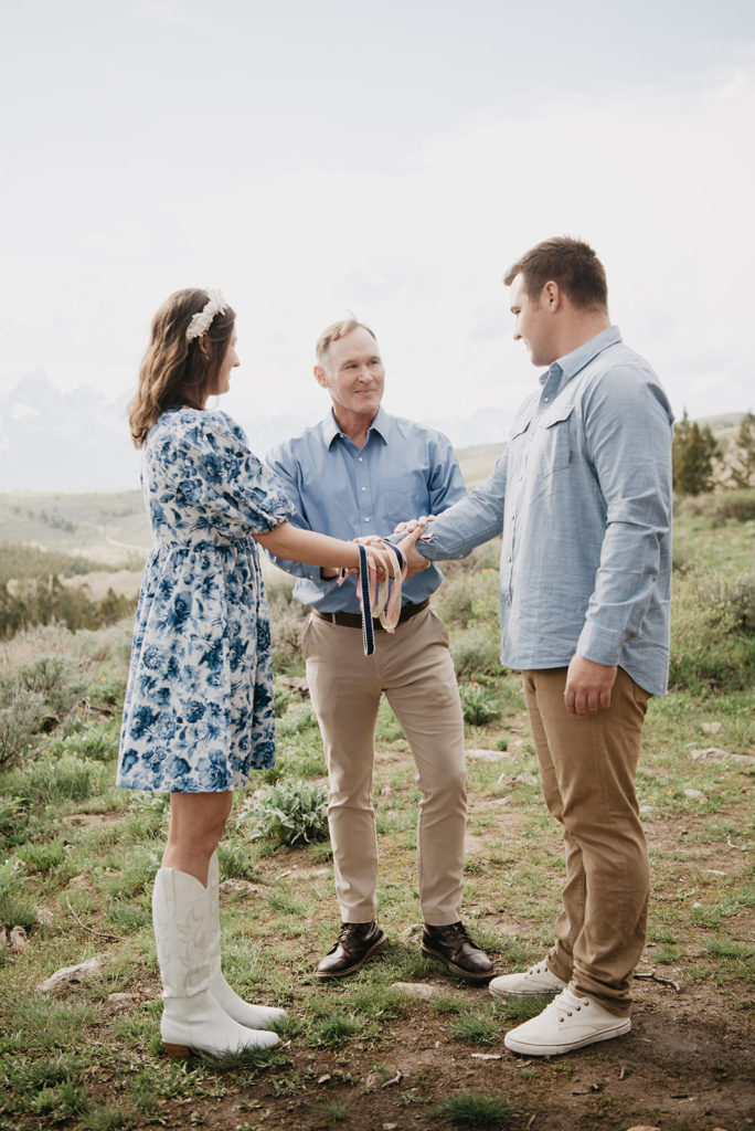 Jackson Hole elopement photographers capture outdoor mountains top wedding ceremony with bride and groom tying the knot with pastel ribbons