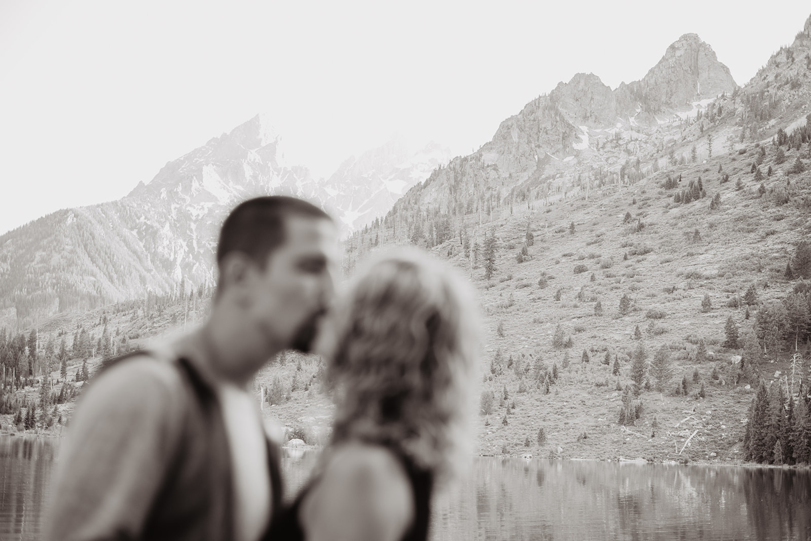 man and woman kissing out of focus with the focus being on the Grand Tetons behind them for their Jackson Hole honeymoon pictures with Jackson Hole wedding photographer