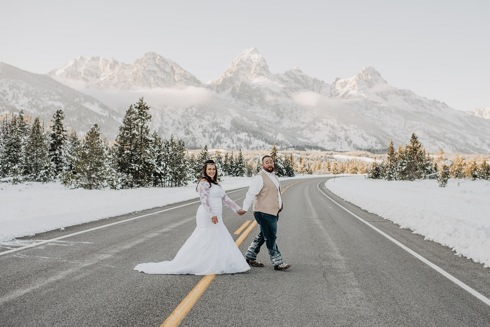 bride and groom holding hands and walking across the road for their winter wedding with the Tetons in the distance with Jackson Hole wedding photographer