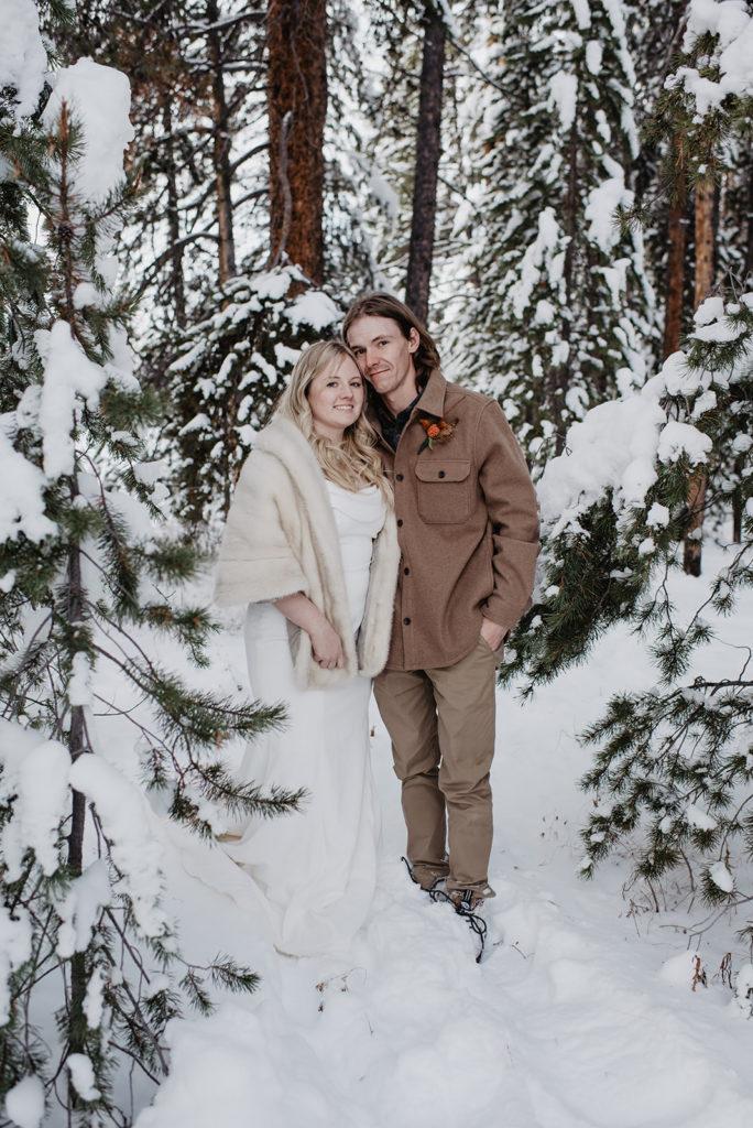 Jackson Hole wedding photographers captures bride and groom standing in the woods that have been recently snowed on for their winter wedding pictures in the Tetons