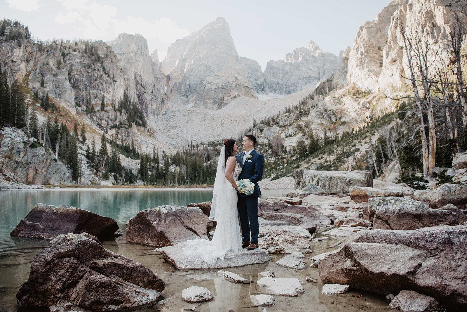 bride and groom standing together at Delta Lake with the Grand Tetons in the distance as they embrace on another for their wedding portraits