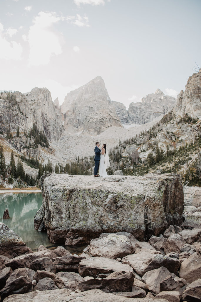 Grand Teton wedding with Jackson Hole photographer capturing bride and groom standing on top of a large boulder in front of a bright blue lake with the Tetons in the distance