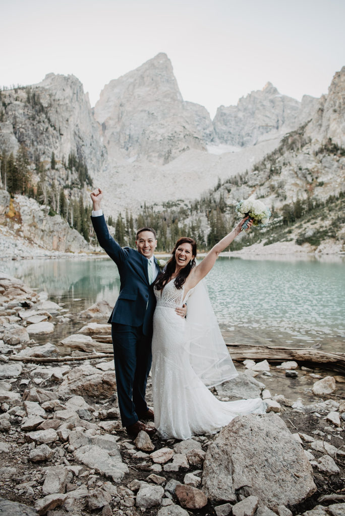 Jackson Hole photographer captures grand teton wedding with bride and groom hugging each other with one arm while their other arm is in the air in celebration in front of Delta Lake  