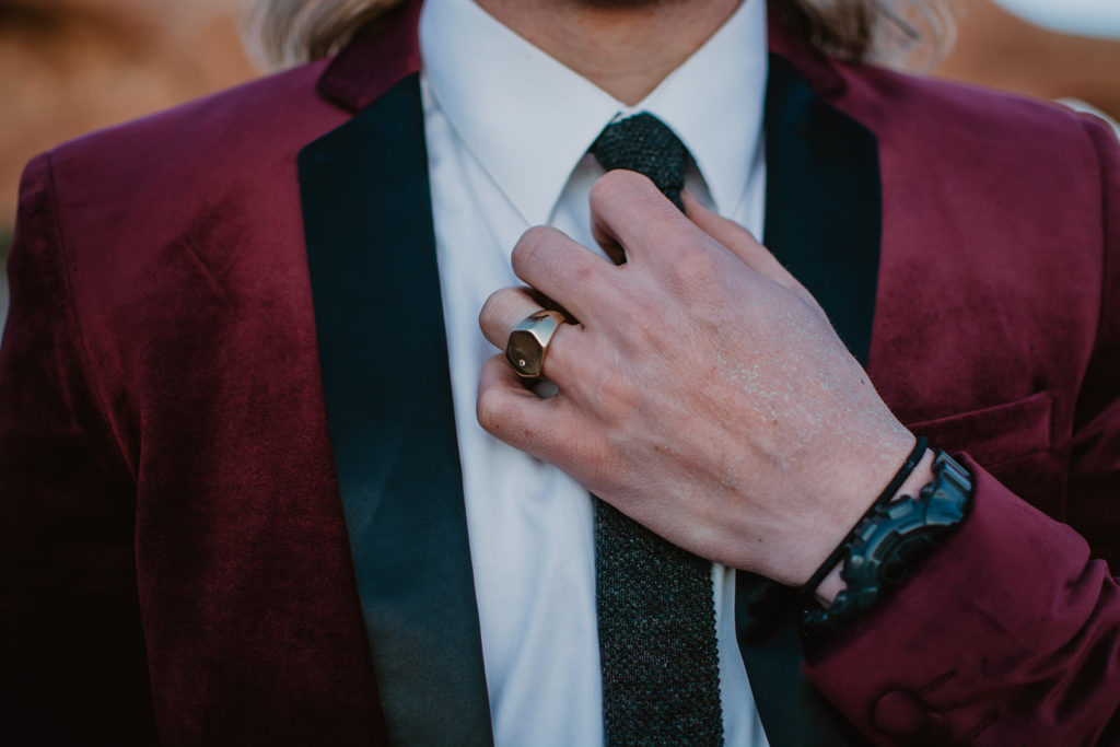 groom adjusting his black tie under his burgundy suit coat showing his wedding ring captured by Utah wedding photographer 