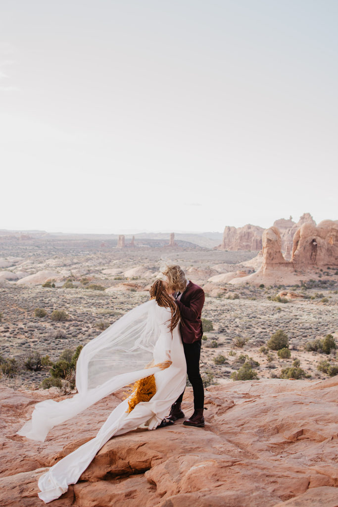 Arches National Park wedding with bride and groom on an outlook on top of red rock with the wind blowing the brides veil as her groom kisses her passionately with a beautiful view in the distance captured by Utah elopement photographer