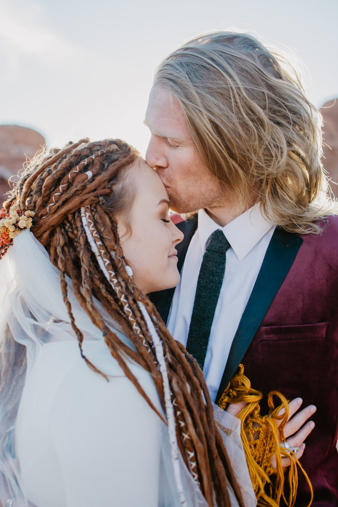 Utah elopement photographer captures groom kissinf his bride on the forehead during their Arches national park  wedding with groom in a burgundy suit and bride wearing an orange shawl 