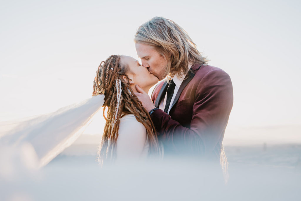 bridal portraits by utah elopement photographer with bride and groom kissing as the sun sets in the distance as her veil flows in the wind