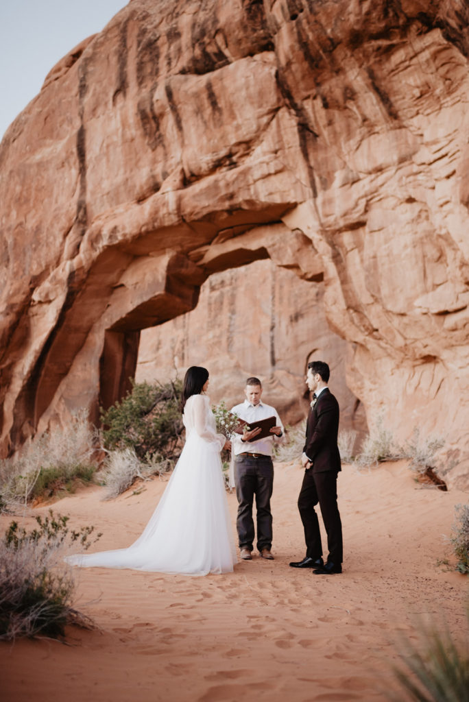 utah elopement photographer captures bride and groom during their outdoor ceremony in Moab's red rocks saying their vows under a large red arch