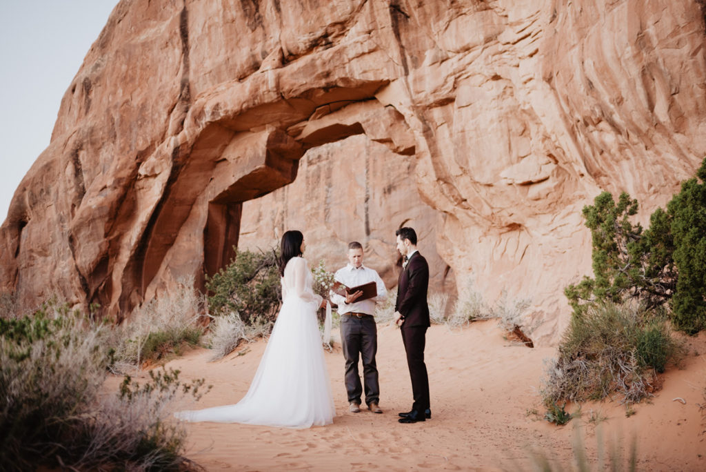 bride and groom standing with their officiant in the red rock of Moab for their utah elopement with a large arch behind them captured by Utah elopement photographer