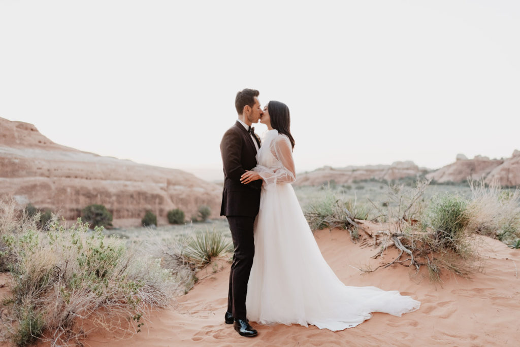 utah elopement photographer captures bride and groom kissing in southern utah at Arches National Park with shrubs and dessert landscape surrounding them while in their luxury wedding attire 