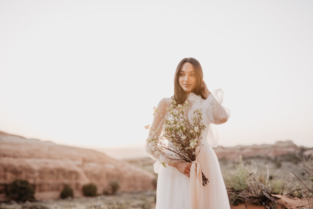 wedding portraits by utah elopement photographer with elegant wedding dress on a dark hair bride holding a wild flower bouquet at sunset for her Moab wedding