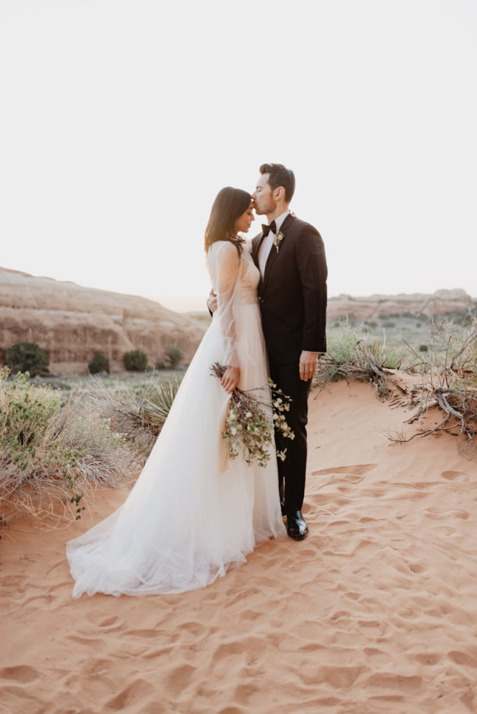 Utah Elopement Photographer captures bride and groom embracing on an outlook in Arches National Park wedding whie the bride holds her wedding bouquet to her side as her groom kisses her forehead