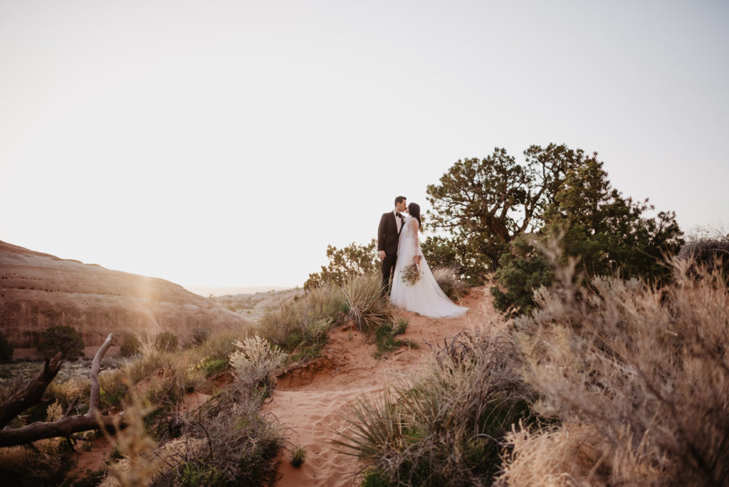 bride and groom standing on top of a hill in the desert of southern utah as the groom leans in to kiss his bride while the sunsets in the distance captured by Utah elopement photographer 